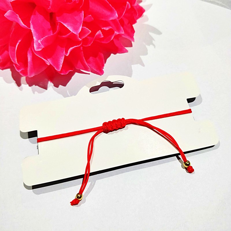 Czerwona bransoletka z cienkiego sznurka jubilerskiego z umieszczoną w centralnym punkcie naturalną, jasną muszelką kauri, zabezpieczoną po bokach dwiema małymi złotymi kuleczkami.