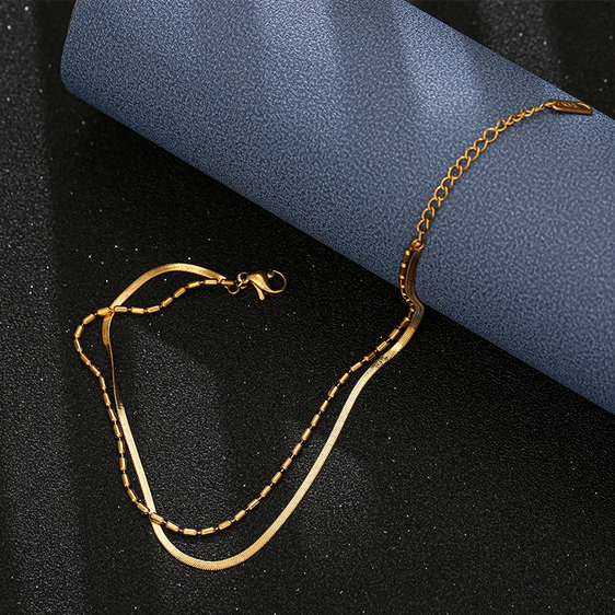 Złota bransoletka na kostkę złożona z dwóch połączonych łańcuszków: gładkiej, płaskiej żmijki oraz drugiego z drobnymi, podłużnymi koralikami.