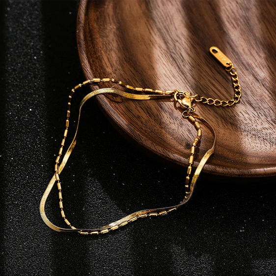 Złota bransoletka na kostkę złożona z dwóch połączonych łańcuszków: gładkiej, płaskiej żmijki oraz drugiego z drobnymi, podłużnymi koralikami.