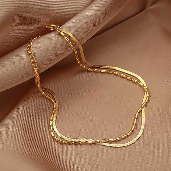 Złota bransoletka na kostkę złożona z dwóch połączonych łańcuszków: gładkiej, płaskiej żmijki oraz drugiego z drobnymi, podłużnymi koralikami.