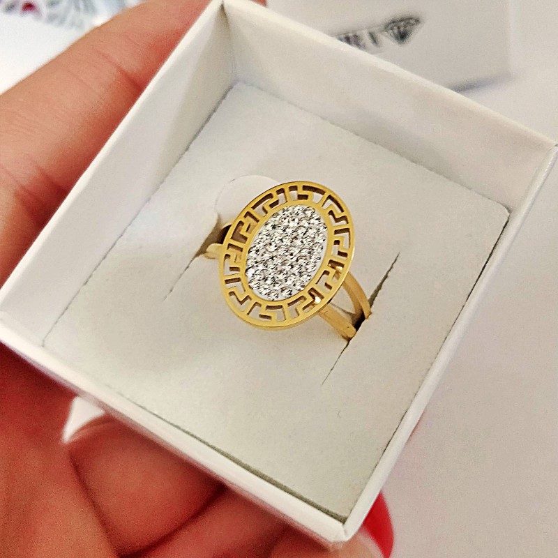 Złoty pierścionek ze stali chirurgicznej z dużą, owalną tarczą, otoczoną ażurowym wzorem greckim i wypełnioną wewnątrz gęsto osadzonymi, błyszczącymi cyrkoniami.