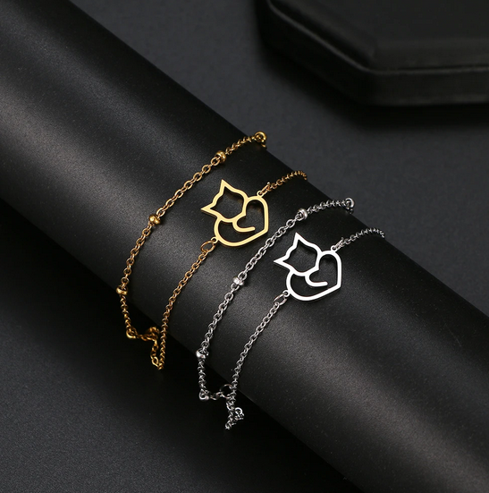 Dwie cienkie bransoletki łańcuszkowe ze stali chirurgicznej (czerwona i srebrna) z minimalistyczną zawieszką w kształcie ażurowego konturu siedzącego kotka z ogonem zawiniętym w serce.