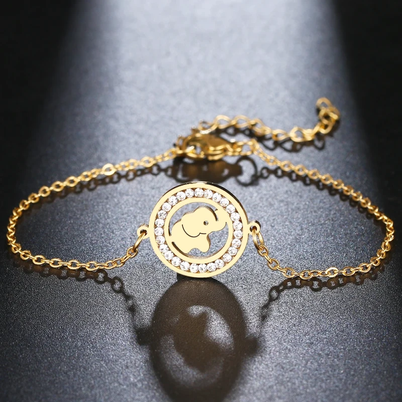 Złota, podwójna bransoletka łańcuszkowa ze stali chirurgicznej. W centrum znajduje się płaska, okrągła zawieszka z wyciętym wewnątrz motywem słonika z uniesioną trąbą, obwiedziona wianuszkiem drobnych, lśniących cyrkonii.