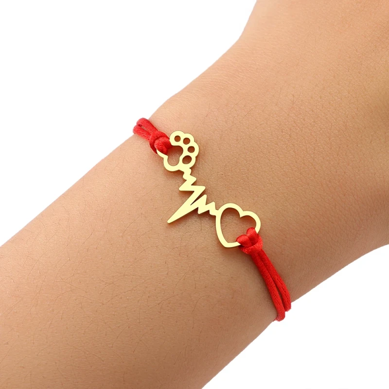 Minimalistyczna bransoletka z czerwonego sznurka jubilerskiego z regulowanymi węzełkami. Na środku znajduje się płaska, złota zawieszka ze stali chirurgicznej łącząca motyw psiej łapki, linii tętna (EKG) oraz serca.