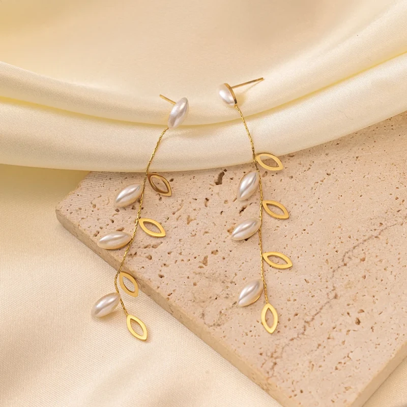 Długie złote kolczyki wiszące ze stali chirurgicznej (7,5 cm) z motywem gałązki, zdobione ażurowymi listkami i białymi podłużnymi perłami.