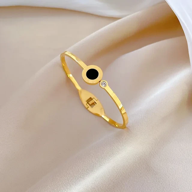 Złota, sztywna bransoletka ze stali chirurgicznej typu bangle, ozdobiona centralnym czarnym kółkiem z wygrawerowanymi cyframi rzymskimi oraz małą, błyszczącą cyrkonią umieszczoną tuż obok.