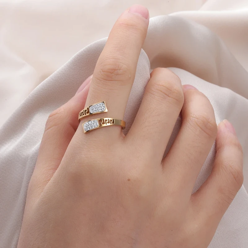 Złoty pierścionek ze stali chirurgicznej o otwartym, spiralnym kształcie. Ozdobiony jest wyciętym wzorem greckim oraz panelami z białą emalią, w której osadzone są jasne cyrkonie.