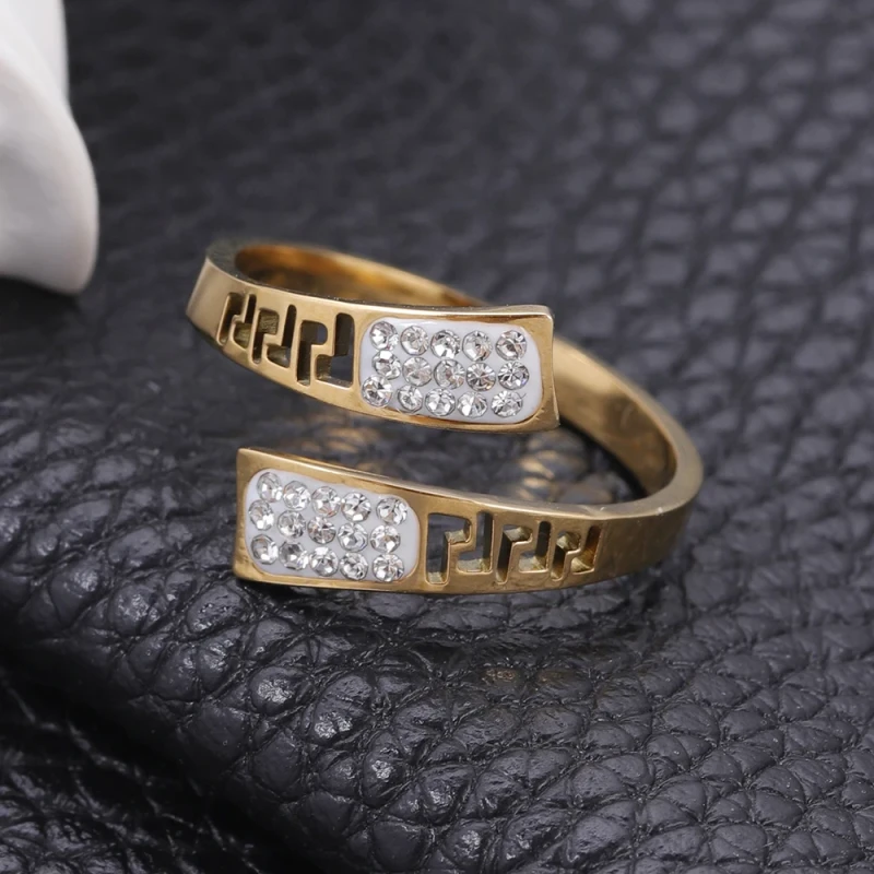 Złoty pierścionek ze stali chirurgicznej o otwartym, spiralnym kształcie. Ozdobiony jest wyciętym wzorem greckim oraz panelami z białą emalią, w której osadzone są jasne cyrkonie.