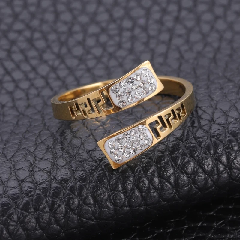 Złoty pierścionek ze stali chirurgicznej o otwartym, spiralnym kształcie. Ozdobiony jest wyciętym wzorem greckim oraz panelami z białą emalią, w której osadzone są jasne cyrkonie.