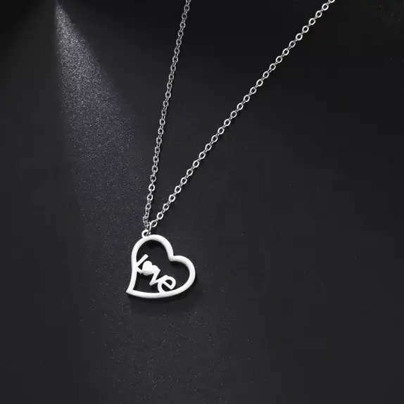 Srebrny naszyjnik ze stali chirurgicznej na ciemnym tle. Zawieszka w kształcie otwartego serca z wkomponowanym w środku napisem LOVE. Zamiast litery O znajduje się małe serduszko.