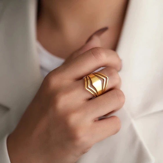 Szeroki, pozłacany pierścionek ze stali chirurgicznej. Charakteryzuje się nowoczesnym, ażurowym wzorem składającym się z grubych, falujących i geometrycznych pasów. Szerokość na froncie ok. 24 mm.
