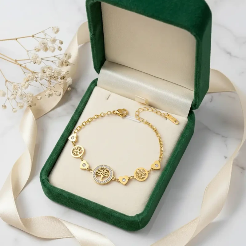 Złota bransoletka łańcuszkowa ze stali chirurgicznej zaprezentowana w zielonym pudełeczku jubilerskim. Posiada okrągłą, centralną zawieszkę z Drzewem Życia otoczoną cyrkoniami oraz mniejsze zawieszki: drzewka i małe serduszka.