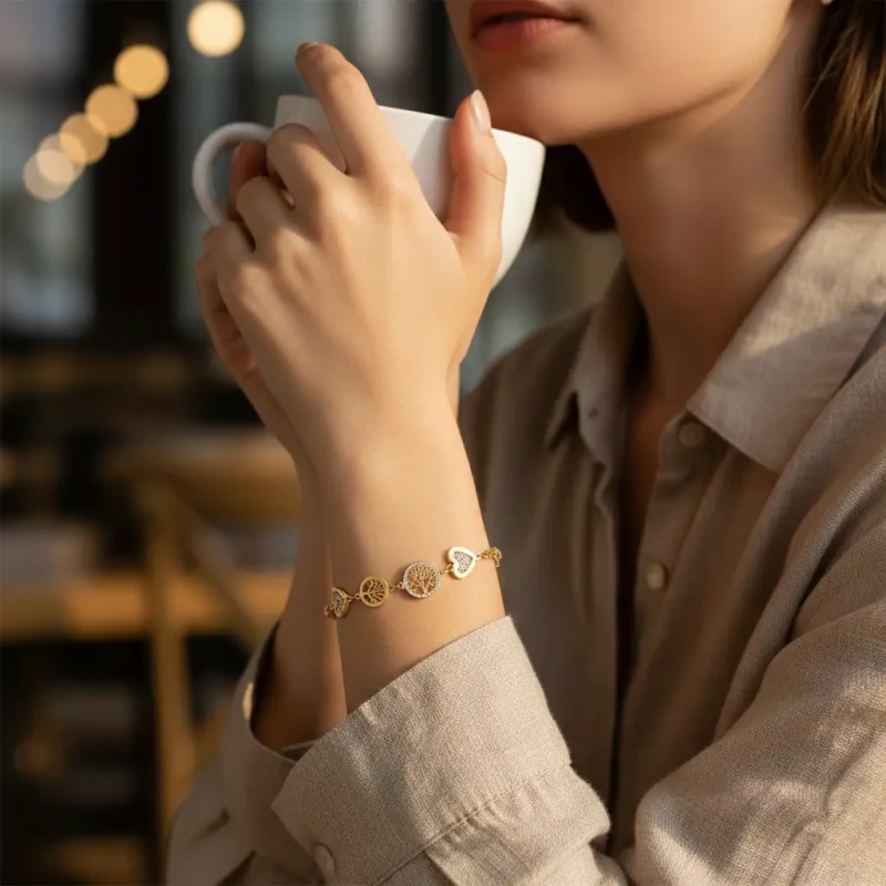 Złota bransoletka łańcuszkowa ze stali chirurgicznej zaprezentowana w zielonym pudełeczku jubilerskim. Posiada okrągłą, centralną zawieszkę z Drzewem Życia otoczoną cyrkoniami oraz mniejsze zawieszki: drzewka i małe serduszka.