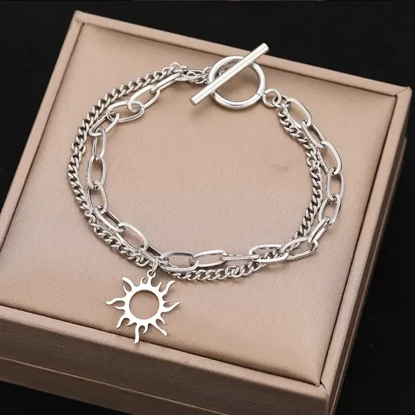 Srebrna, podwójna bransoletka ze stali chirurgicznej na kobiecym nadgarstku. Składa się z dwóch łańcuszków: gęstej pancerki oraz splotu typu spinacz z doczepioną ażurową zawieszką w kształcie słońca.