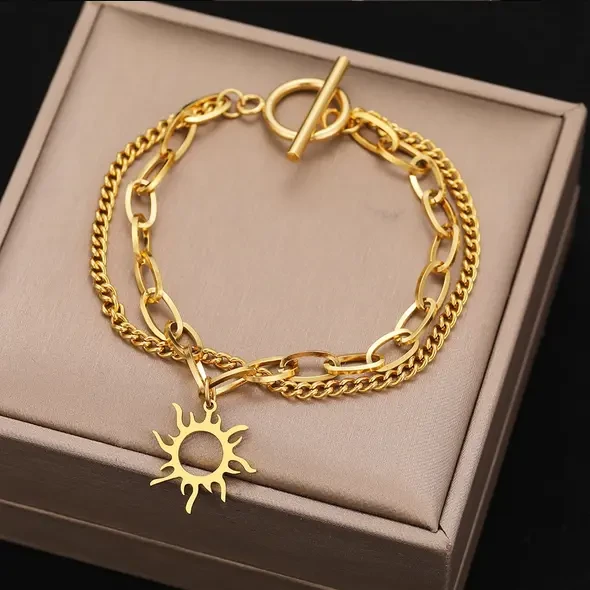 Złota, podwójna bransoletka ze stali chirurgicznej na kobiecym nadgarstku. Składa się z dwóch łańcuszków: gęstej pancerki oraz splotu typu spinacz z doczepioną ażurową zawieszką w kształcie słońca.