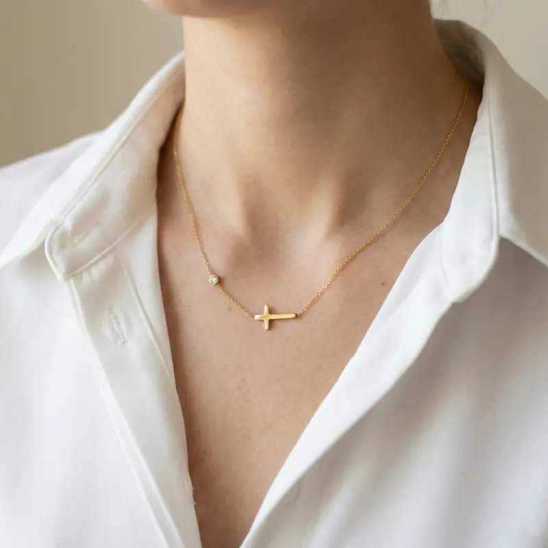 Złoty naszyjnik celebrytka ze stali chirurgicznej na szyi, z motywem gładkiego poziomego krzyżyka oraz pojedynczą cyrkonią na łańcuszku.