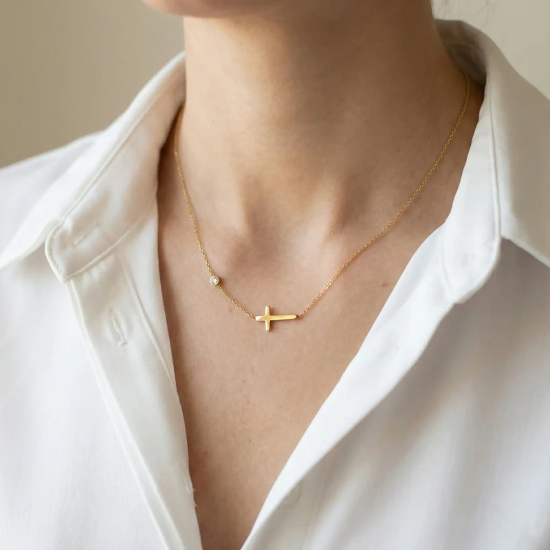 Złoty naszyjnik celebrytka ze stali chirurgicznej na szyi, z motywem gładkiego poziomego krzyżyka oraz pojedynczą cyrkonią na łańcuszku.