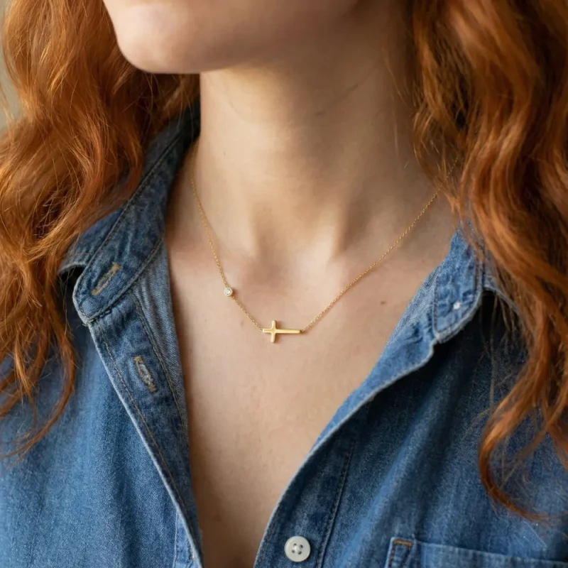 Złoty naszyjnik celebrytka ze stali chirurgicznej na szyi, z motywem gładkiego poziomego krzyżyka oraz pojedynczą cyrkonią na łańcuszku.