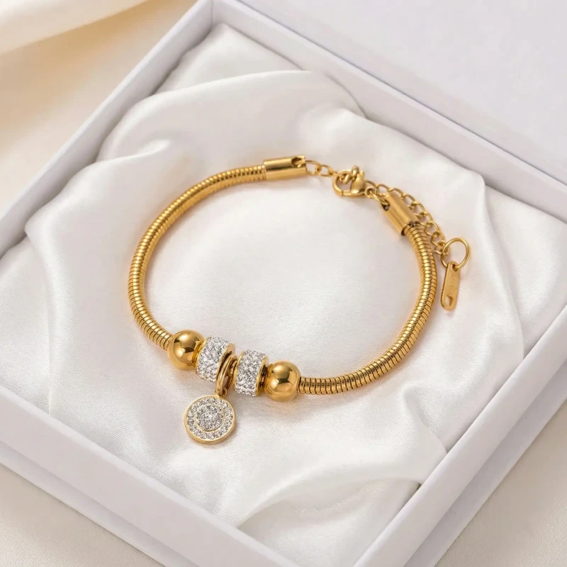 Złota bransoletka ze stali chirurgicznej o gładkim splocie typu pandorka (żmijka). W jej centralnej części znajduje się ozdobne koło gęsto wysadzane drobnymi, jasnymi cyrkoniami.