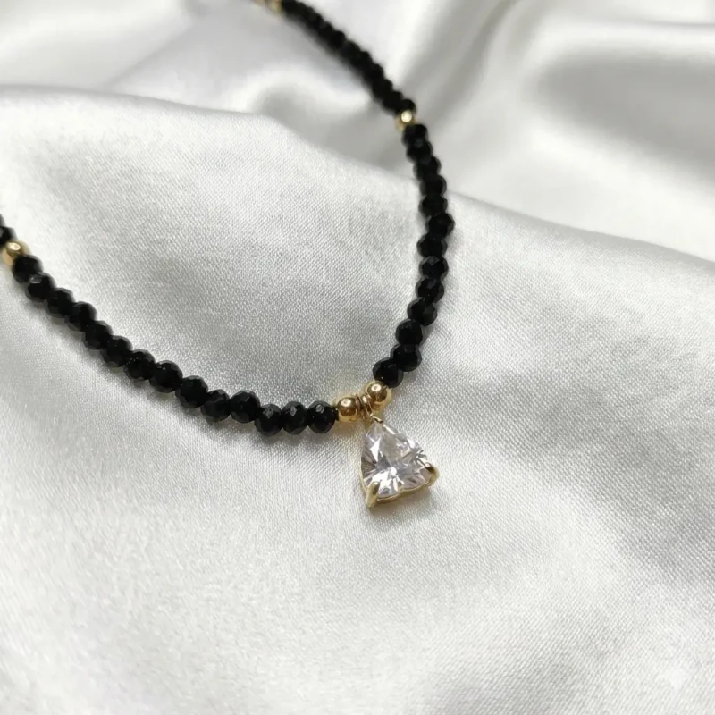 Delikatny naszyjnik z drobnych czarnych koralików ze złotymi akcentami i kryształową zawieszką w kształcie trójkąta, prezentowany na szyi.