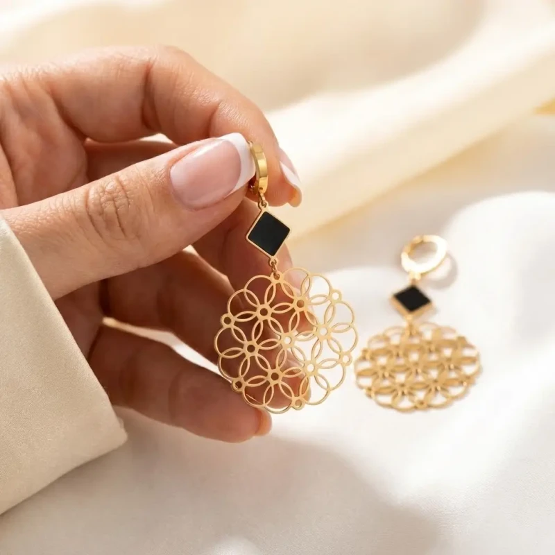 Długie złote kolczyki ze stali chirurgicznej z czarnym rombem u góry i dużą, ażurową rozetą o wzorze kwiatowym na dole.