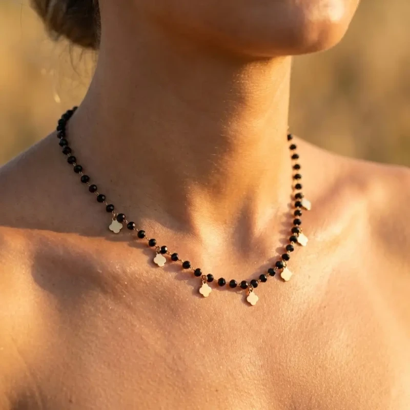 Złoty naszyjnik ze stali chirurgicznej złożony z czarnych fasetowanych koralików oraz wiszących złotych blaszek w kształcie koniczynek, prezentowany na szyi.