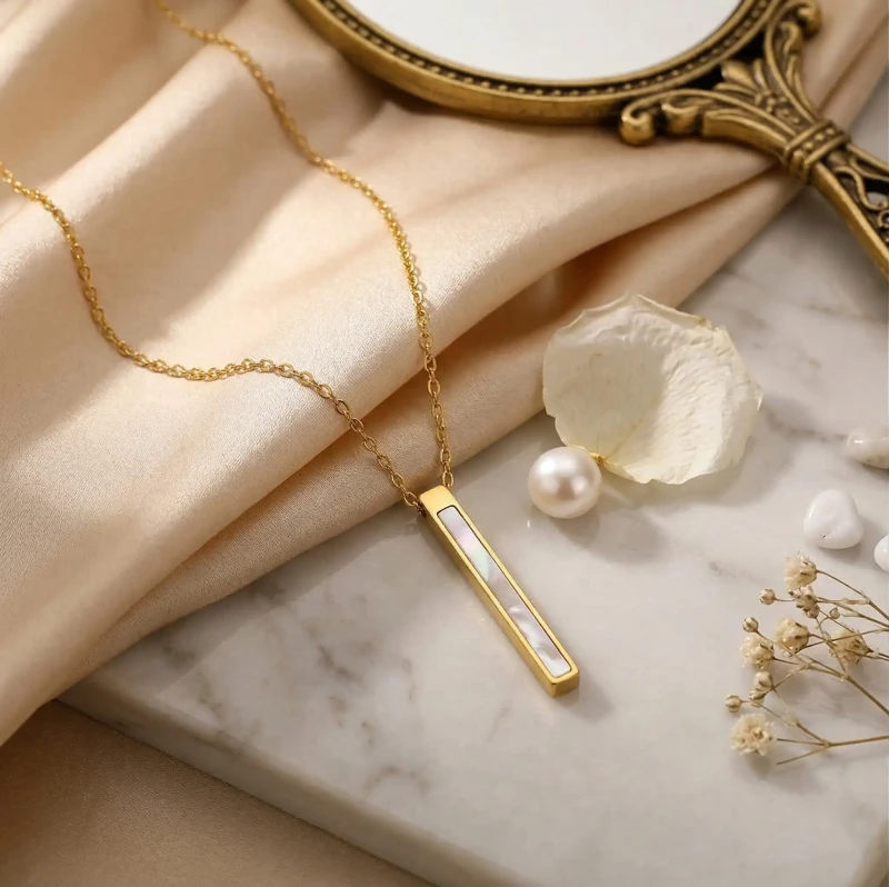 Złoty naszyjnik ze stali chirurgicznej z pionową, prostokątną zawieszką w kształcie pałki wypełnionej lśniącą masą perłową, zaprezentowany w luksusowej aranżacji na atłasowym materiale.