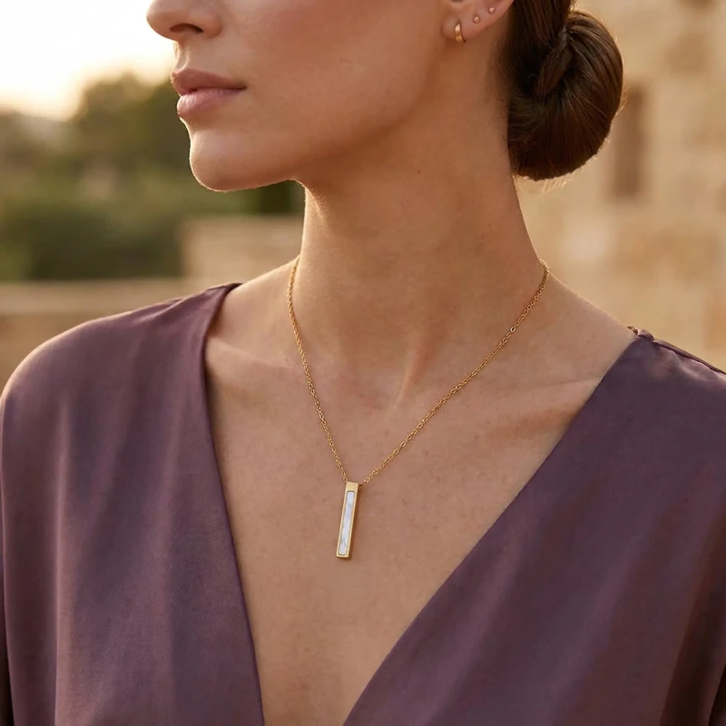 Złoty naszyjnik ze stali chirurgicznej z pionową, prostokątną zawieszką w kształcie pałki wypełnionej lśniącą masą perłową, zaprezentowany w luksusowej aranżacji na atłasowym materiale.