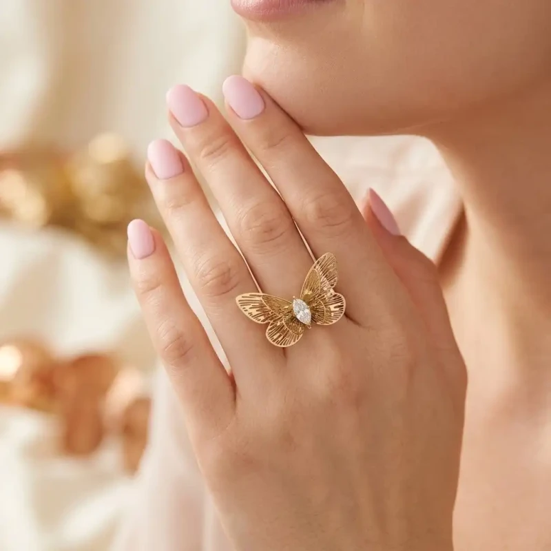 Złoty pierścionek typu statement ze stali chirurgicznej. Pierścionek ma kształt okazałego, rozłożystego motyla z kunsztownymi, ażurowymi wycięciami na skrzydłach oraz centralnie osadzoną cyrkonią w kształcie markizy (łezki).