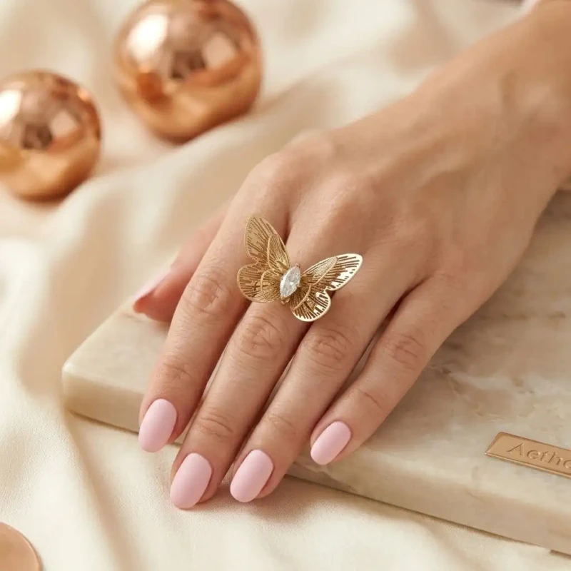 Złoty pierścionek typu statement ze stali chirurgicznej. Pierścionek ma kształt okazałego, rozłożystego motyla z kunsztownymi, ażurowymi wycięciami na skrzydłach oraz centralnie osadzoną cyrkonią w kształcie markizy (łezki).