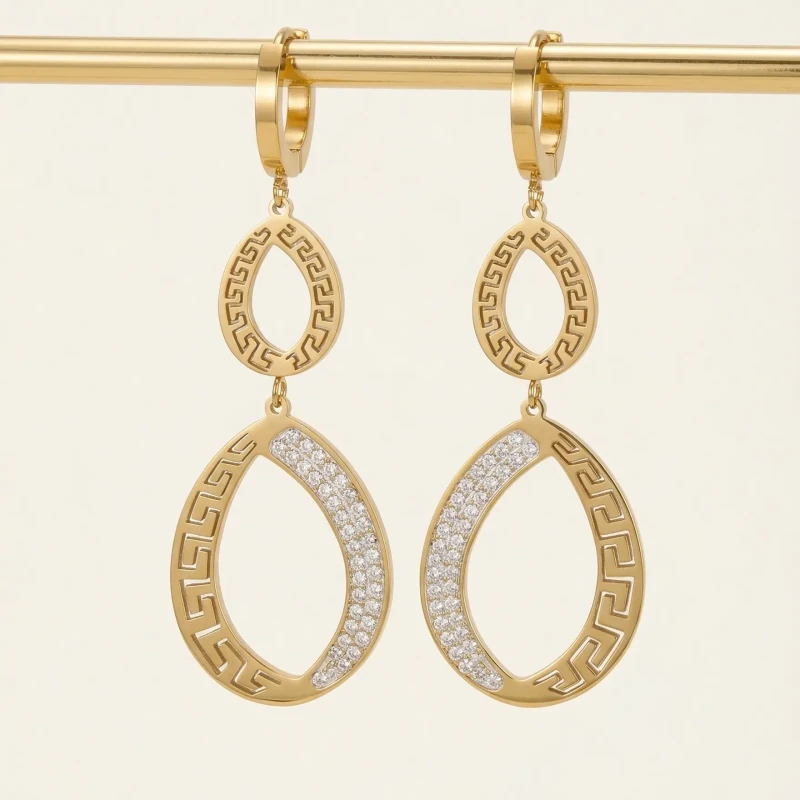 Długie, złote kolczyki wiszące ze stali chirurgicznej na sztyfcie. Kształt podwójnych łez ozdobionych antycznym wzorem greckim i lśniącymi cyrkoniami.