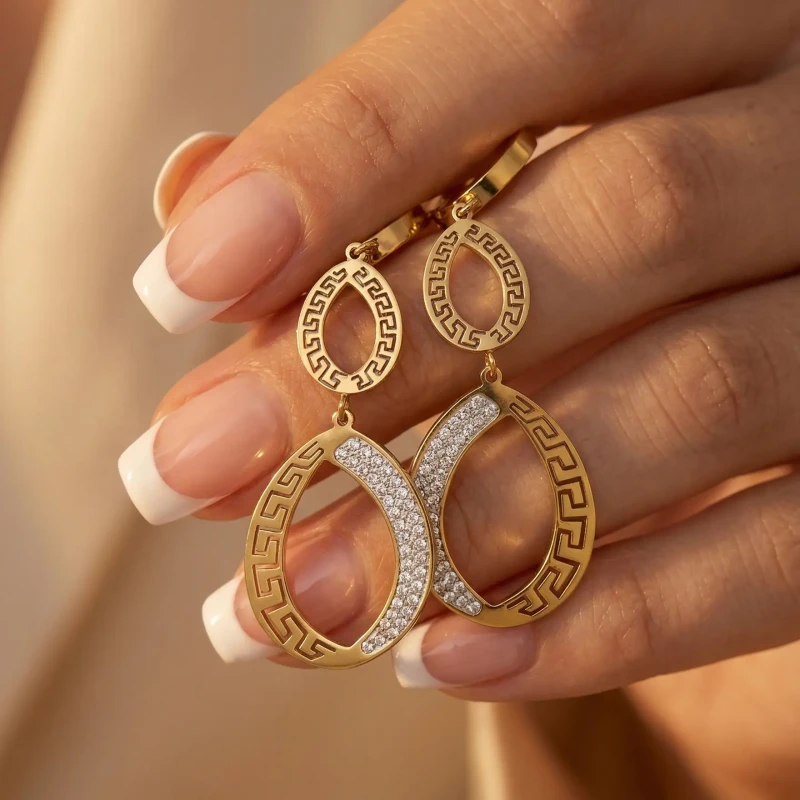 Długie, złote kolczyki wiszące ze stali chirurgicznej na sztyfcie. Kształt podwójnych łez ozdobionych antycznym wzorem greckim i lśniącymi cyrkoniami.