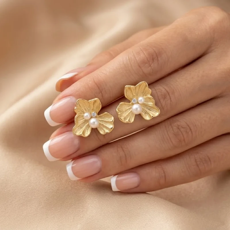 Złote kolczyki na sztyfcie w kształcie trójpłatkowego kwiatu o ryflowanej strukturze, zdobione dwiema perłami i małą cyrkonią.