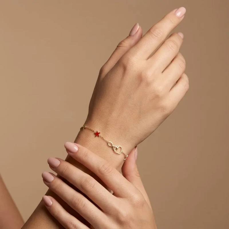 Złota bransoletka celebrytka na nadgarstku. Na cienkim łańcuszku znajduje się zawieszka w kształcie otwartego serca złączonego ze znakiem nieskończoności, a po bokach umieszczono małe gwiazdki..webp