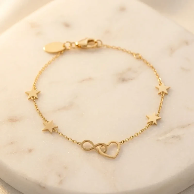 Złota bransoletka celebrytka na nadgarstku. Na cienkim łańcuszku znajduje się zawieszka w kształcie otwartego serca złączonego ze znakiem nieskończoności, a po bokach umieszczono małe gwiazdki...webp
