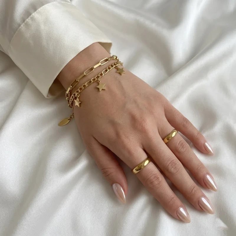 Złota, podwójna bransoletka ze stali chirurgicznej. Łączy łańcuszek o prostokątnym splocie spinaczowym z drugim łańcuszkiem ozdobionym wiszącymi, płaskimi gwiazdkami.