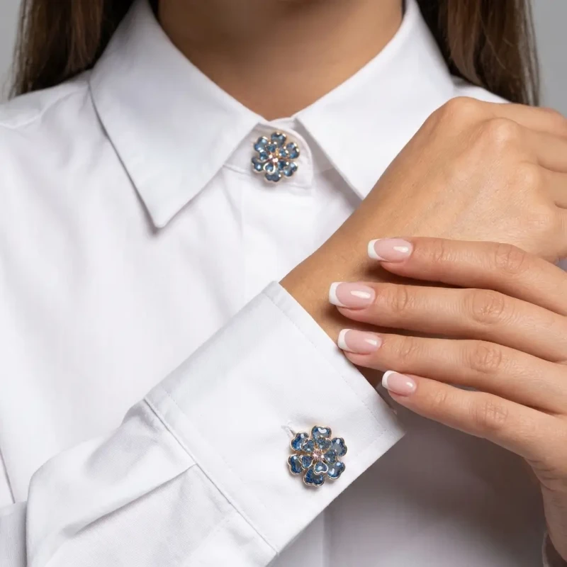 Komplet 2 niebiesko-złotych nakładek na guziki (3,5 cm). Mają kształt wielowarstwowego kwiatu z fasetowanych, błękitnych kryształków w złotej oprawie. Pokazano zapięcie na mankietach białej koszuli.