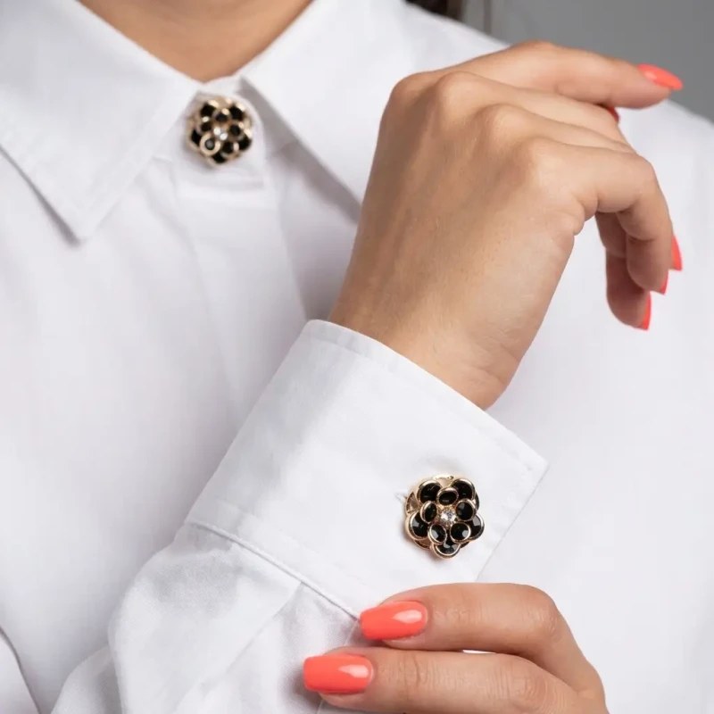 Komplet 2 czarno-złotych nakładek na guziki (3,5 cm). Mają kształt eleganckiego kwiatu z zaokrąglonymi, czarnymi płatkami w złotej oprawie i cyrkonią w centrum. Pokazano zapięcie na koszuli.