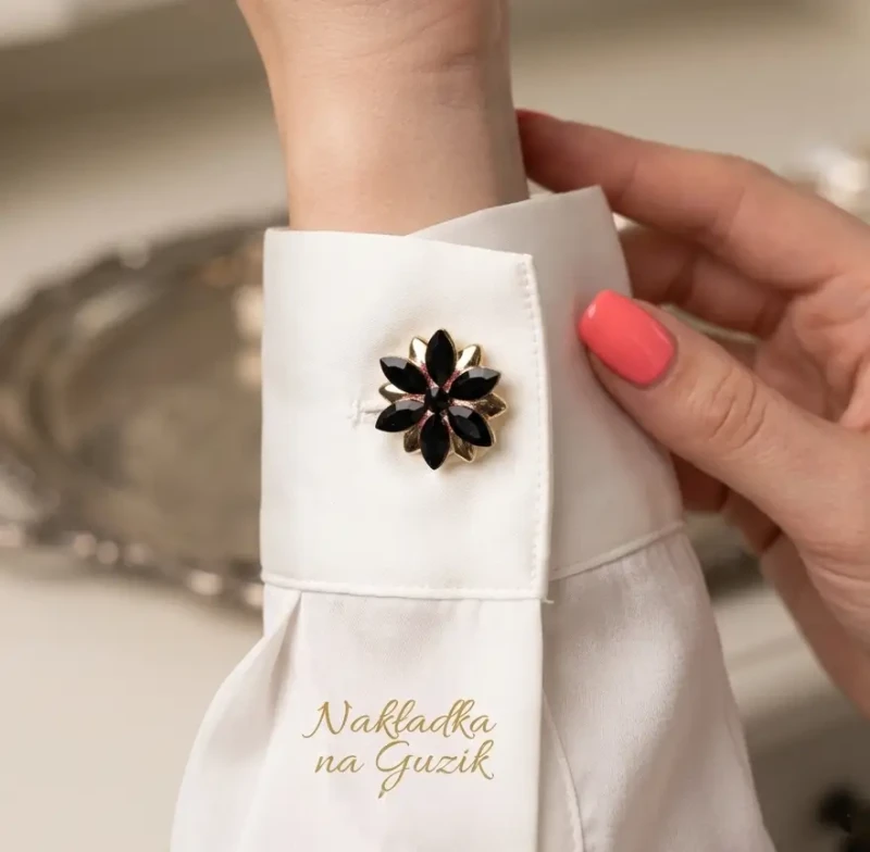 Komplet 2 czarno-złotych nakładek na guziki (3,5 cm). Kształt eleganckiego kwiatu ułożonego z ostro zakończonych, czarnych kamieni w złotej oprawie. Pokazano zapięcie na mankiecie białej, jedwabnej koszuli.