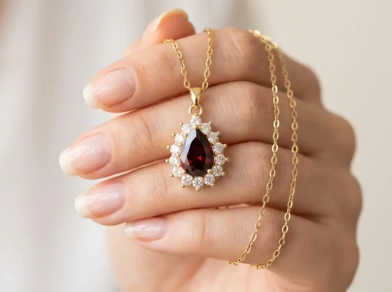Elegancki, pozłacany naszyjnik ze stali chirurgicznej. Na delikatnym łańcuszku zawieszono duży, fasetowany kryształ w kształcie kropli (łezki) w głębokim, rubinowym (czerwonym) kolorze